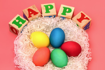 Five Easter colored eggs lie in a nest on top of cubes with the words happy on a gently pink background. Horizontal photo.