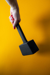 hand of a young woman holding a black wooden hammer, nails covered with red varnish, bright yellow background, close-up, vertical shot