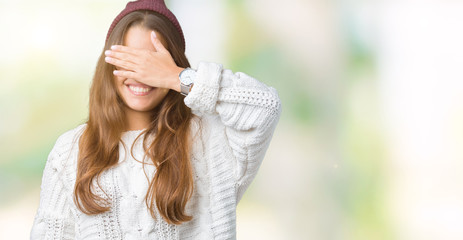 Young beautiful brunette hipster woman wearing glasses and winter hat over isolated background smiling and laughing with hand on face covering eyes for surprise. Blind concept.