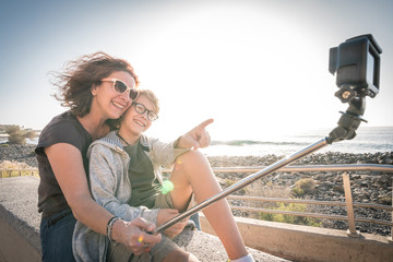 Smiling and happy mom takes selfie with her little blond kid with action cam Mother with son take vacation selfie photo with sea background Woman with the young man takes beautiful cell kiss pictures