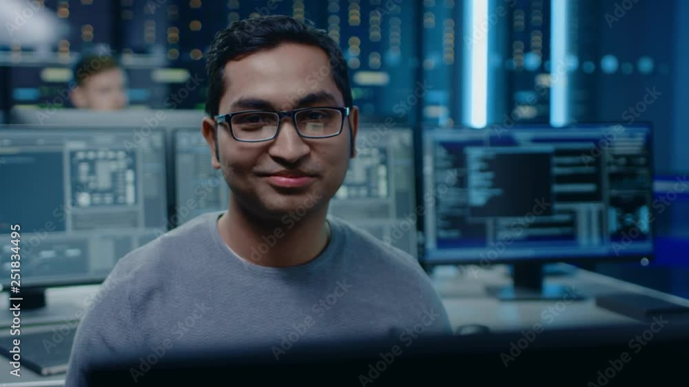 Poster Portrait of a Smart and Handsome Software Developer Wearing Glasses Working on a Personal Computer, Smiles at the Camera. In the Background Unfocused Personal Computers with Screens in Technical Room
