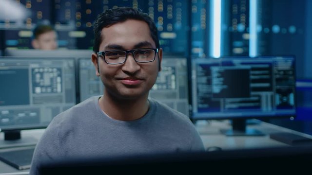 Portrait of a Smart and Handsome Software Developer Wearing Glasses Working on a Personal Computer, Smiles at the Camera. In the Background Unfocused Personal Computers with Screens in Technical Room
