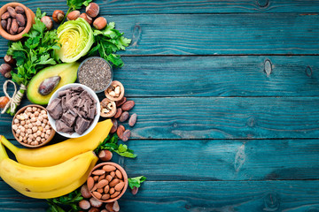 Foods containing natural magnesium. Mg: Chocolate, banana, cocoa, nuts, avocados, broccoli, almonds. Top view. On a blue wooden background. - obrazy, fototapety, plakaty