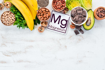 Foods containing natural magnesium. Mg: Chocolate, banana, cocoa, nuts, avocados, broccoli, almonds. Top view. On a white wooden background. - obrazy, fototapety, plakaty
