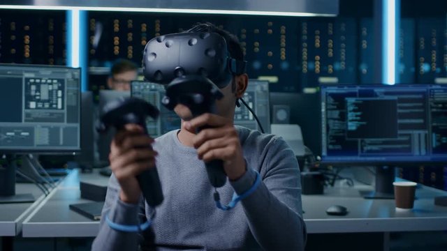 Portrait of Software Delevoper Wearing Virtual Reality Headset Using Controllers to Develop and Program VR Gaming and Applications. In the Background Technology Developing Studio with Computers