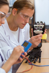 Close up of young male scientist, focusing on work, holding mechanical hand. Handsome man wearing white uniform and protective glasses, wiping with yellow cloth inside prosthesis at laboratory.