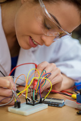 Close up of female engineering student working in laboratory, using voltage and current tester. Cheerful girl wearing in white uniform and protective glasses, inspect parts that using in project.