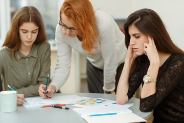 Pupils hardly thinking under difficult tasks during classes. Young teacher helping and explaining graphics and schematics to her students. Red haired tutor checking homeworks of beautiful girls.