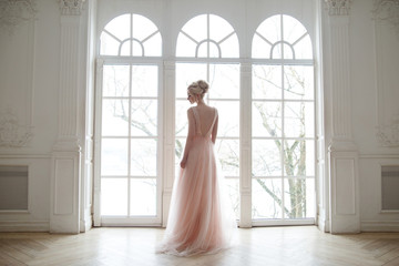 The bride in a chic lush pink dress stands near the window in the beautiful hall.