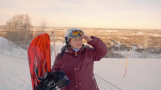 Sporty senior ederly female woman holds snowboard outdoors snowy background. Woman looking far sideward. Active lifestyle winter vacation