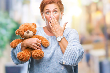 Senior caucasian woman holding teddy bear over isolated background cover mouth with hand shocked...