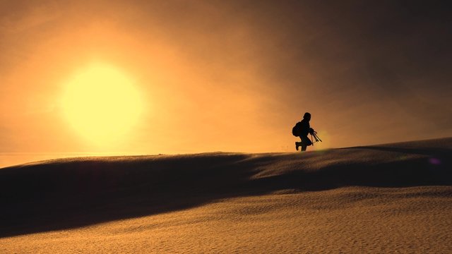 photographer traveler in winter goes on snowy ridge in rays of yellow sunset. mountaineer with camera and tripod is walking in snow along top of holom. © zoteva87