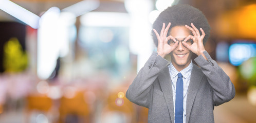 Young african american business man with afro hair wearing glasses doing ok gesture like binoculars sticking tongue out, eyes looking through fingers. Crazy expression.