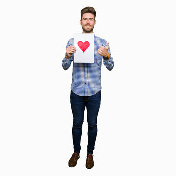 Young handsome man holding paper with red heart happy with big smile doing ok sign, thumb up with fingers, excellent sign