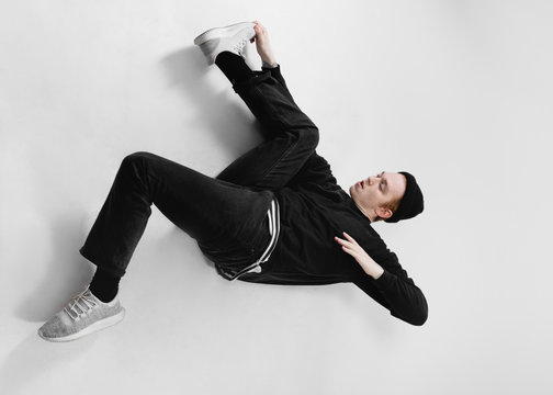 Freestyle dancer dressed in black jeans, sweatshirt, hat and gray sneakers is dancing lying on the floor in the studio on the white background