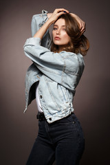 Fototapeta premium Beautiful brunette girl with long flowing hair dressed in jeans jacket and jeans poses holding her hands on her head on the dark background in the studio