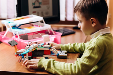 Boy dressed in green sweater sits at the desk with computer and looks at the robot that he made...