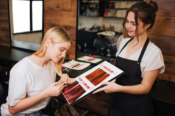 Young beautiful woman and hairdresser comparing her blonde hair with color hair from palette before...