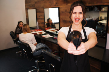 Fototapeta na wymiar Ready to work Emotion beautiful woman hairdresser with black apron looking at camera while holding professional hair dryer on salon background. Beauty and people concept