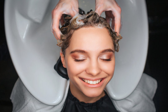 Top view of happy female customer smiling with closed eyes while hairdresser massaging hair. Young woman relaxing in beauty salon