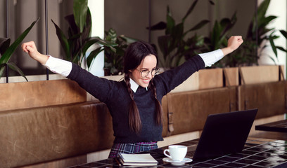 Schoolgirl with braids rejoycing with raising hands while studying with laptop