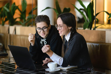 Two schoolgirls in spectacles having positive conversation while using laptop computer on cafe interior