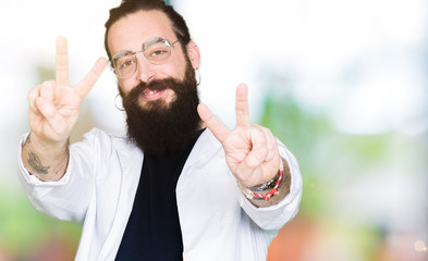 Doctor therapist man with long hair and bear wearing white coat smiling looking to the camera showing fingers doing victory sign. Number two.