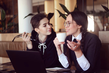 Two cheerful classmates drinking coffee and talking with each other while studying at the cafe
