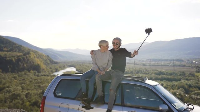 Happy couple sitting on roof of car and possing on the smart phone camera in the mountain.