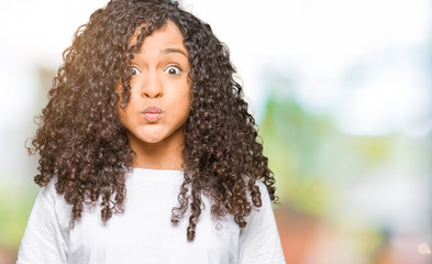 Young beautiful woman with curly hair wearing white t-shirt puffing cheeks with funny face. Mouth inflated with air, crazy expression.