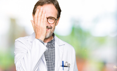 Middle age doctor men wearing medical coat covering one eye with hand with confident smile on face and surprise emotion.