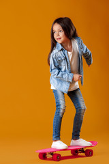 Stylish little child girl with skateboard in jeans clothes on orange background