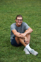 Cheerful young male enjoy free time in public park while sitting on grass. Attractive student boy smiling and looking at camera.