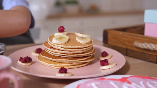 Plate of delicious pancakes decorated for festive breakfast with pieces of banana and raspberries on kitchen table. Little boy's hand decorating holiday breakfast for Mother's day.