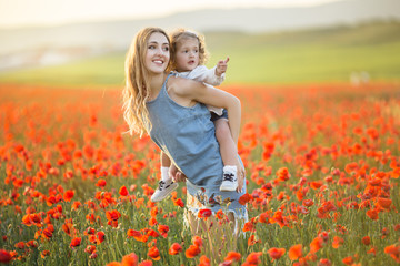 Beautiful smiling child girl with mother are having fun in field of red poppy flowers over sunset...