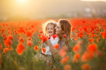 Beautiful smiling baby girl with mother are having fun in field of red poppy flowers over sunset lights, spring time