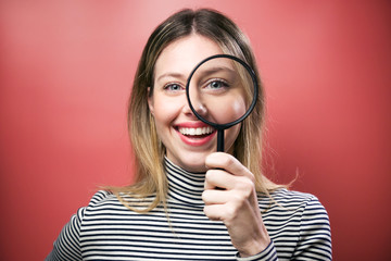 Funny young woman looking through magnifying glass at the camera over pink background. - obrazy, fototapety, plakaty