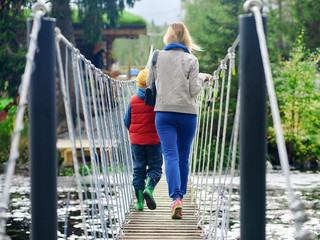 Mom and son are enjoying their hike. They are in the picturesque nature reserve and moving along the rope bridge across the river.