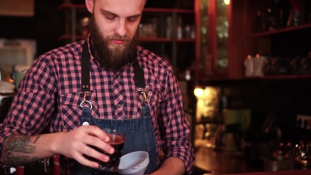 Experienced male barista with tattoos and beard busy making alcohol cocktail on bar counter in slow motion