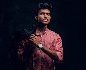 Vogue, fashion, style. Handsome young Indian guy wearing a pink shirt holding a jacket on his shoulder and looking sideways. Studio photo against a dark textured wall