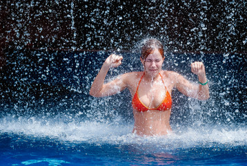laughing and happy girl in an orange swimsuit plays jumping and having fun under a waterfall in the pool