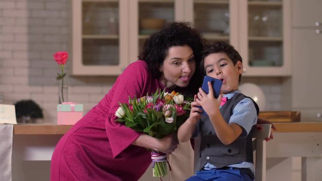 Mother with tulip flowers and cute mixed race preadolescent son holding smart phone grimacing for selfie in kitchen. Family having fun on holiday making amusing faces taking pictures on mobile phone
