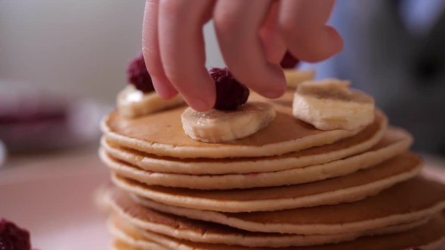 Close up heap of tasty homemade pancakes cooked by loving son for holiday breakfast. Child's hand carefully decorating festive meal with fresh raspberries and bananas for Mother's day