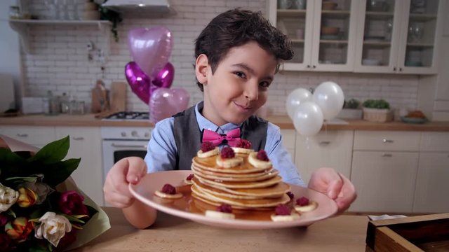 Closeup smiling mixed race son holding plate with pancakes, festively decorated with fruit. Portrait of joyful boy showing delicious breakfast to camera offering it to mother on Mother's day