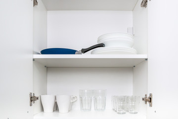Opened kitchen cabinet with plates, glasses, cups and frying pan. Close up.