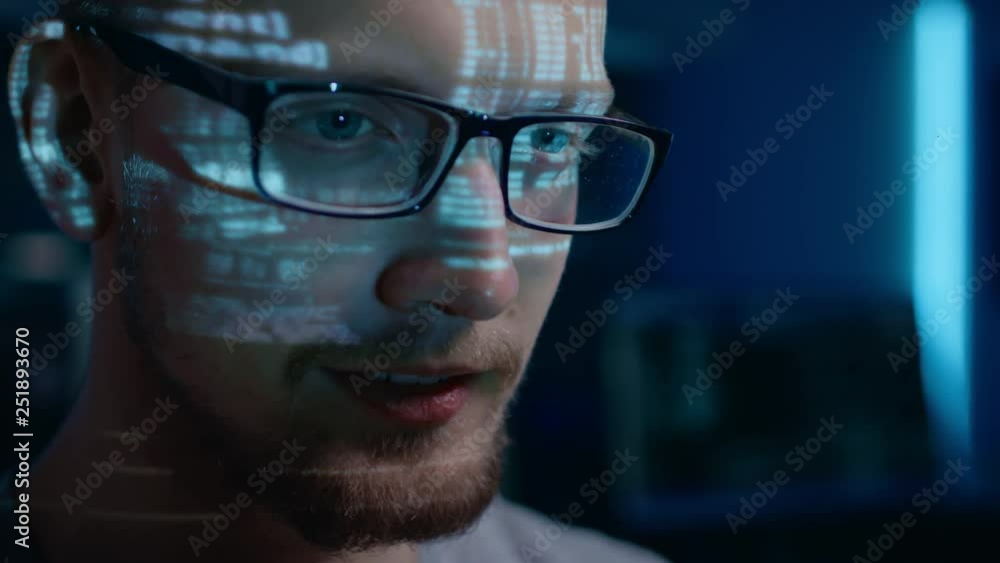 Poster Portrait of Software Developer / Hacker wearing Glasses Working on Computer, Projected Code Numbers and Characters Reflect on His Face. Dark Room Full of Technology. Zoom in Shot