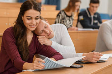 Beautiful woman sitting at table in lecture hall and reading notes while male student sleeping near in morning. Adults in process of studying at university. Concept boring lesson and learning.