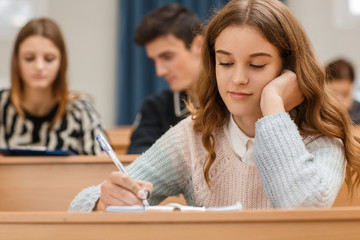 Front view of pretty female student writing material in notebook during lesson. Attractive girl enjoying interesting lecture and getting new knowledge. Concept of studying process.