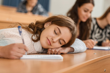 Lazy female student lying on table in lecture hall, keeping pen in hand and sleeping during lesson...