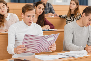 Shocked male student in white sweater with open mouth keeping test and looking at bad mark. Young boy sitting in lecture hall at university and confused with result. Concept of disappointment.